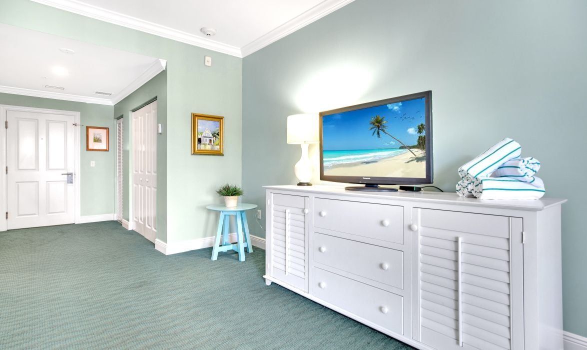 A room with a television on a white dresser, a small blue stool, and a framed picture on the wall.