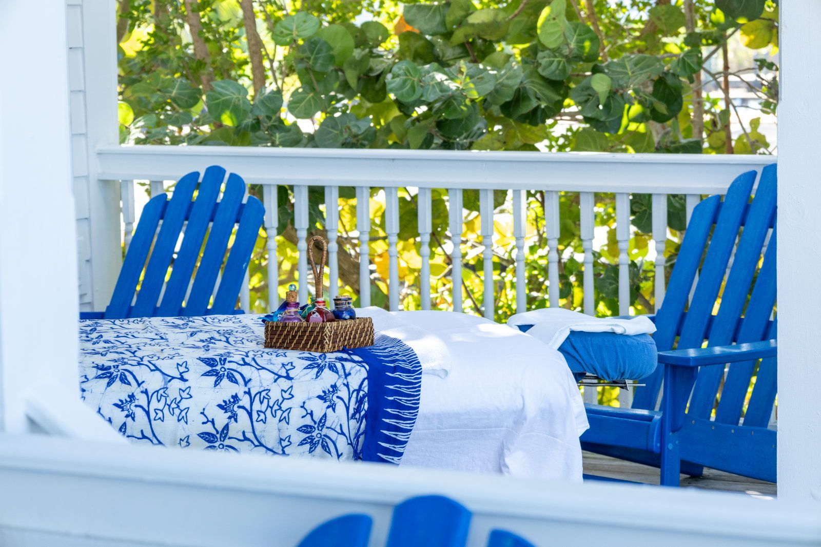 A porch with a white railing, blue chairs, and a bed with blue and white bedding.