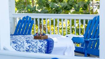 A porch with a white railing, blue chairs, and a bed with blue and white bedding.