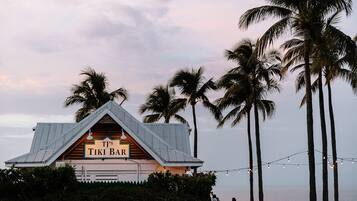 A tiki bar with a sloped roof, surrounded by palm trees and overlooking a body of water.