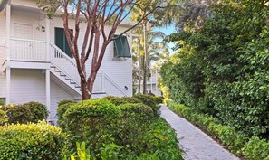 A white building with a staircase leading to a balcony, surrounded by lush greenery and a paved walkway.