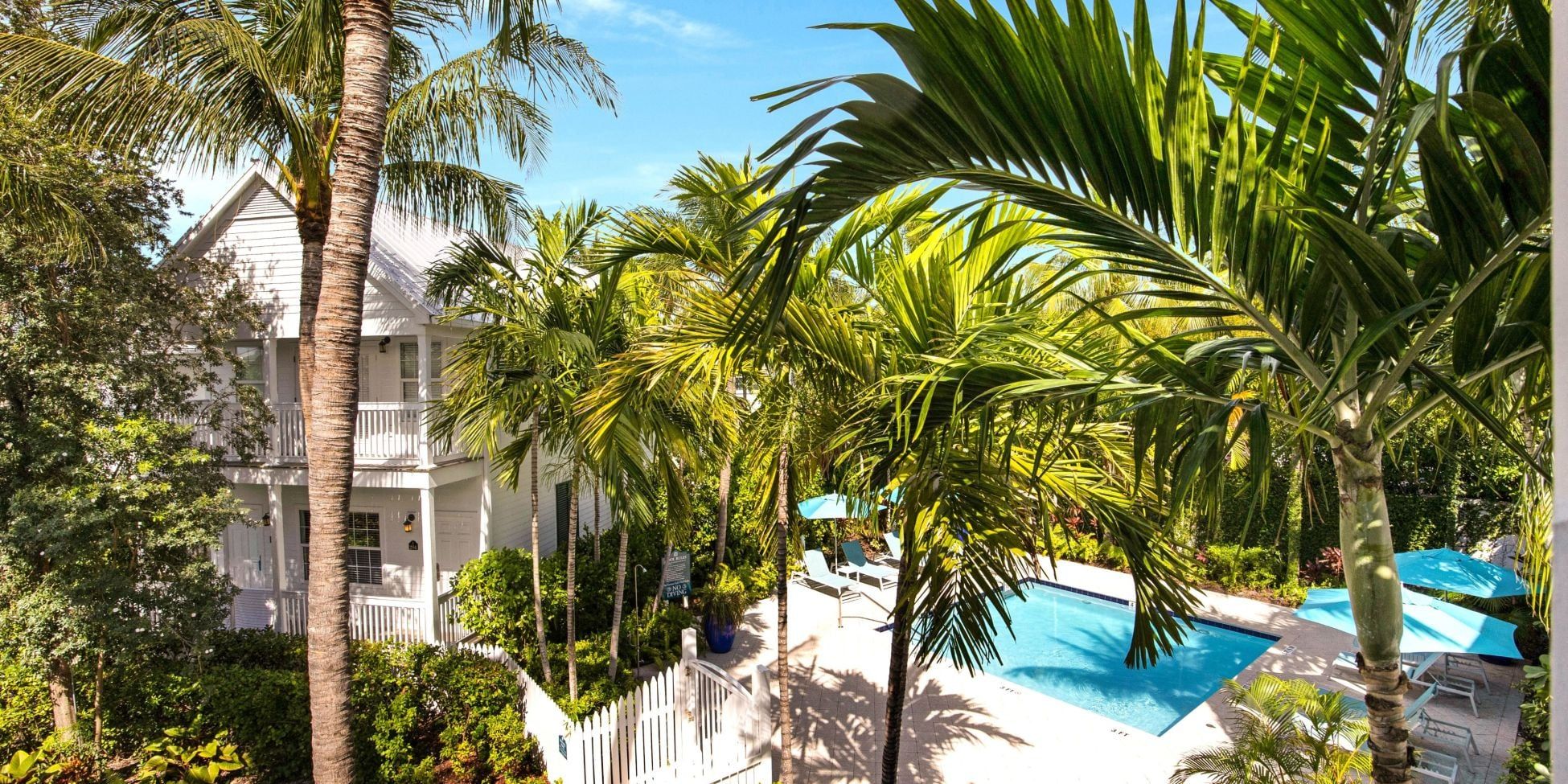 A pool area with lounge chairs and palm trees, surrounded by a white fence and a building with balconies.
