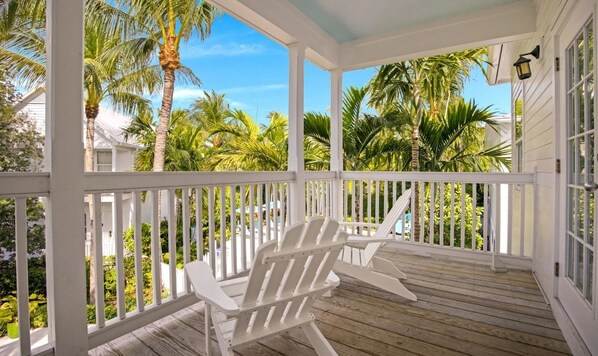 Un porche cubierto con barandillas blancas y suelo de madera, con vistas a un jardín tropical con palmeras.