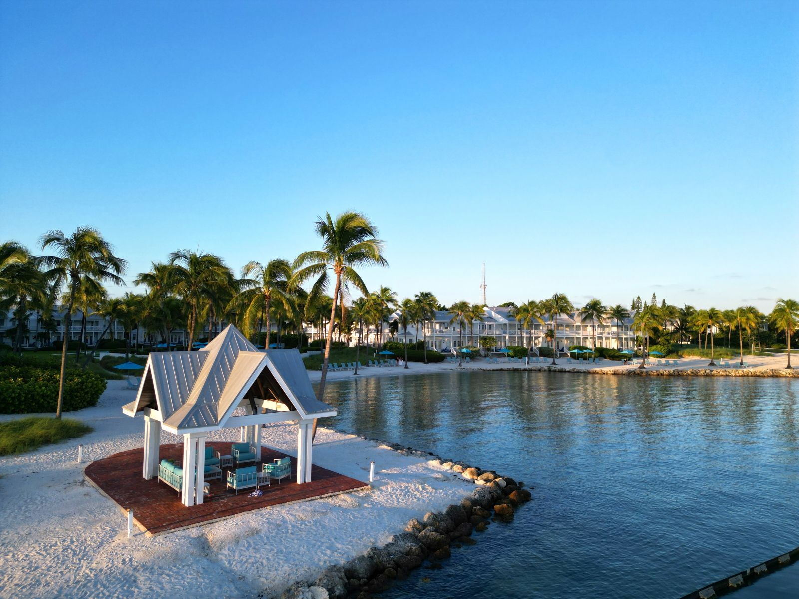 A beachside resort with a small pavilion, palm trees, and a calm lake.