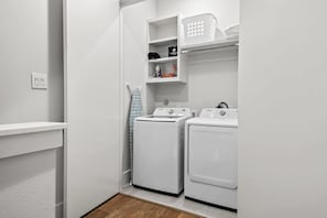 A laundry room with two white washing machines, a drying rack, and a shelf with a bag and a bottle.
