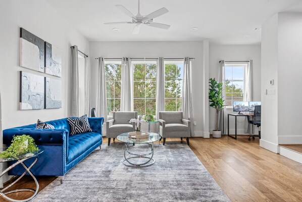 Ein geräumiges Wohnzimmer mit einem blauen Sofa, zwei Sesseln, einem Glastisch und Blick ins Freie durch große Fenster.
