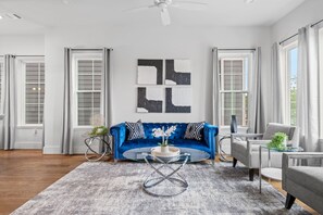 Ein Wohnzimmer mit einem blauen Sofa, einem Glastisch und einem grauen Sessel. Zwei Fenster mit Vorhängen, ein Deckenventilator und eine Wand mit abstrakter Kunst.
