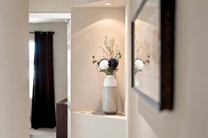 A hallway with a white vase containing flowers on a shelf, a dark curtain, and a framed mirror reflecting the flowers. - Seva Retreat | Gulf Access | Pool |  Game Room (Cape Coral)