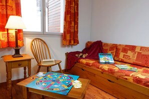 Ein gemütliches Zimmer mit einem Holzstuhl, einem kleinen Tisch mit einem Gesellschaftsspiel, einer Couch mit Decke und Stofftier sowie einem Fenster mit Vorhängen.