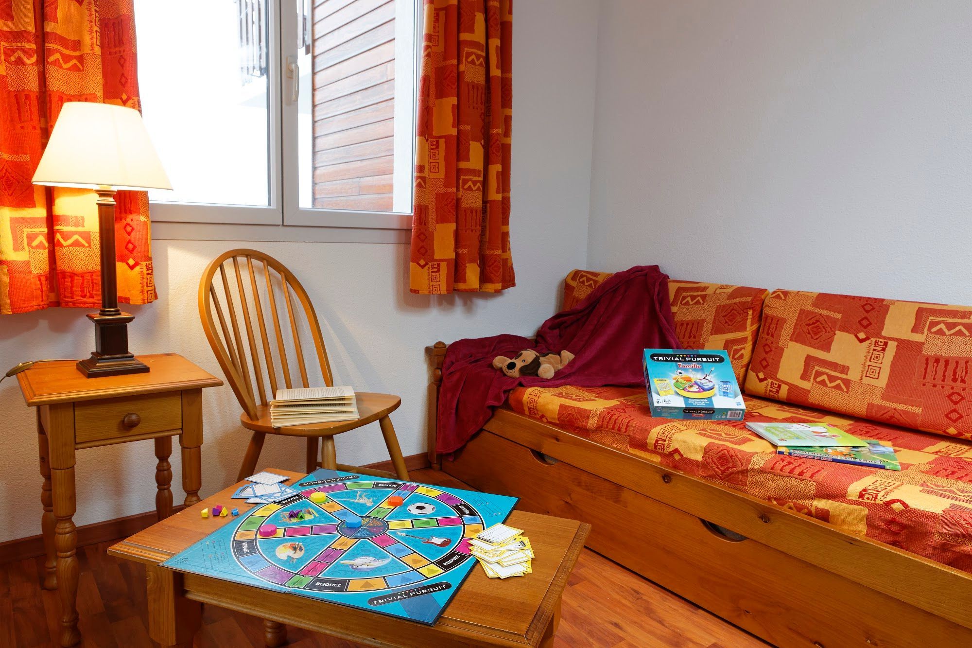 Une pièce chaleureuse avec une chaise en bois, une petite table sur laquelle est posé un jeu de société, un canapé avec une couverture et un jouet en peluche, et une fenêtre avec des rideaux.