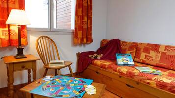 Une pièce chaleureuse avec une chaise en bois, une petite table sur laquelle est posé un jeu de société, un canapé avec une couverture et un jouet en peluche, et une fenêtre avec des rideaux.