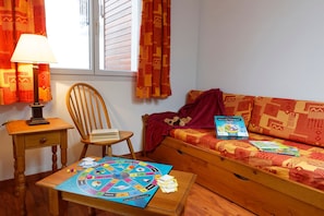 Une pièce chaleureuse avec une chaise en bois, une petite table sur laquelle est posé un jeu de société, un canapé avec une couverture et un jouet en peluche, et une fenêtre avec des rideaux.