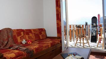 A room with a sofa, a table with a deck of cards, and a view of mountains through a balcony.