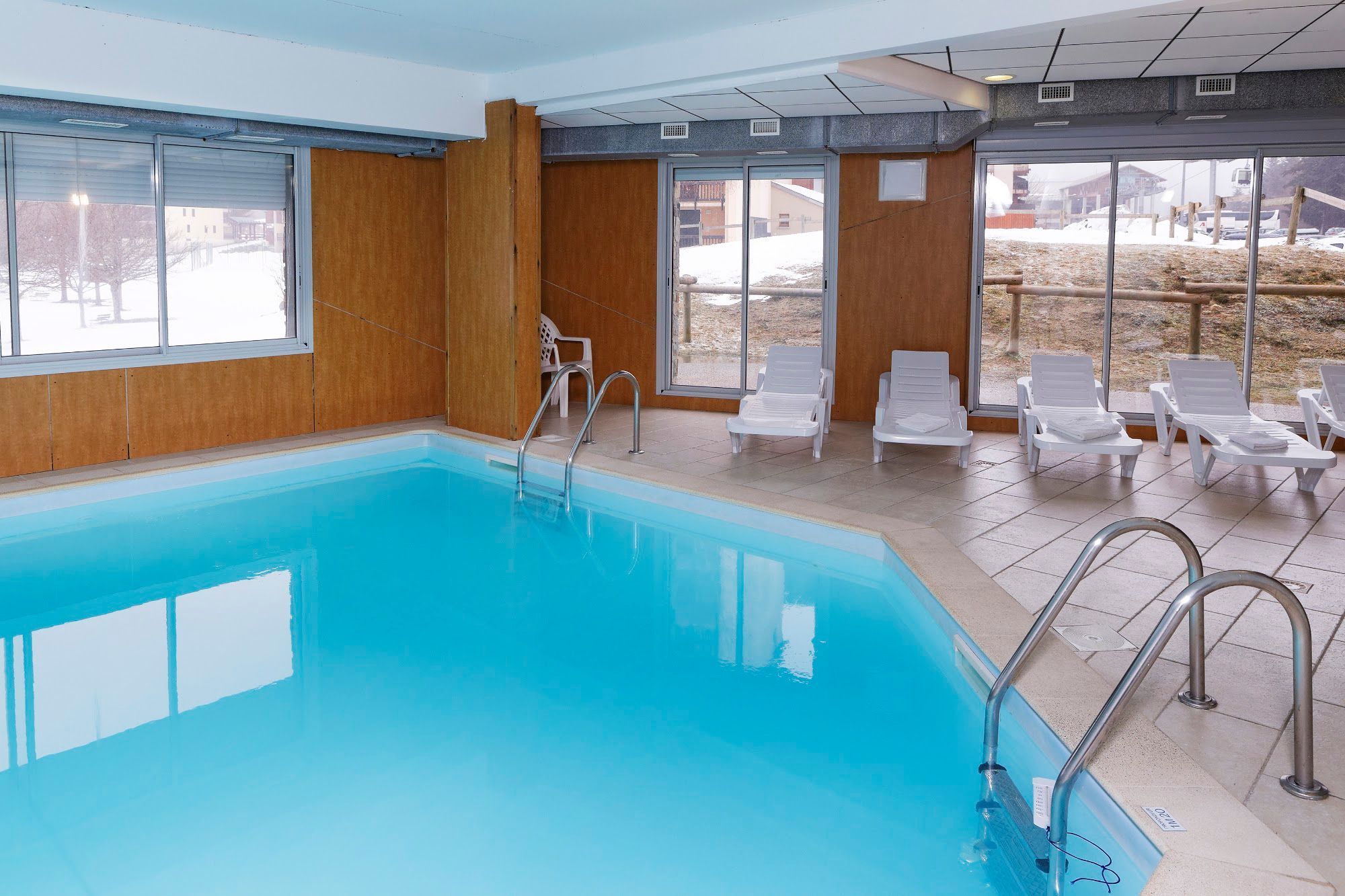 An indoor swimming pool with clear glass walls, a metal ladder, and white lounge chairs.