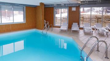 An indoor swimming pool with clear glass walls, a metal ladder, and white lounge chairs.