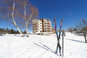 A snow-covered landscape with a multi-story building featuring balconies, and a pair of skis leaning against a pole.