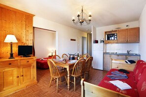 A cozy living room with a dining area, wooden furniture, a television, and a kitchen with a microwave. - À 50m des pistes et du centre | Piscine Chauffée (La Toussuire)