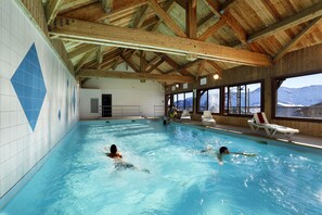 Una piscina interior con vigas de madera, paredes de azulejos y vistas a montañas a través de amplios ventanales.