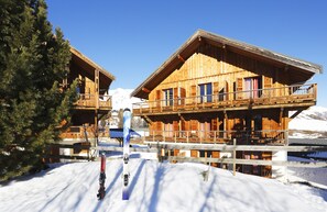 Una cabaña de madera con balcón, equipo de esquí y una montaña nevada al fondo.