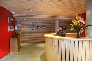 A reception desk with a wooden finish, a vase with flowers, and a red wall with framed pictures. - À 200 m des commerces | Sauna et Bain à remous (Les Contamines-Montjoie)