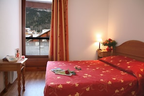 A hotel room with a bed, a desk with a book and a candle, a nightstand with a lamp and flowers, and a window with curtains. - À 200 m des commerces | Sauna et Bain à remous (Les Contamines-Montjoie)