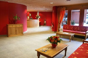 Eine Hotellobby mit einer Rezeption, einer Couch, einem Holztisch mit Blumenvase und einer Zimmerpflanze.