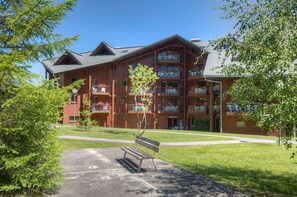 En bygning med flere etager, altaner, et belagt område med en bænk og en klar himmel.