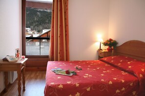 Et hotelværelse med en seng, et skrivebord med en bog og et stearinlys, et natbord med en lampe og blomster, samt et vindue med gardiner.