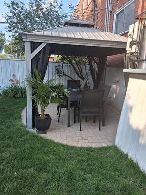 Een overdekte patio met een tafel en stoelen, omgeven door een bakstenen muur en een metalen hek.