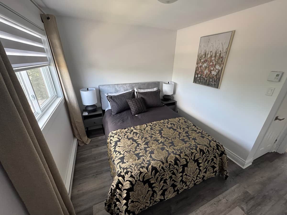 Une chambre à coucher comprenant un lit, des tables de chevet, une commode, une fenêtre avec des rideaux et un luminaire fixé au mur.