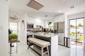 A modern kitchen with white cabinetry, a central island, and a view of a garden through a sliding glass door. - 5 Bed - 3 Full Bath - pool home - 10 mins to beach (Pompano Beach)