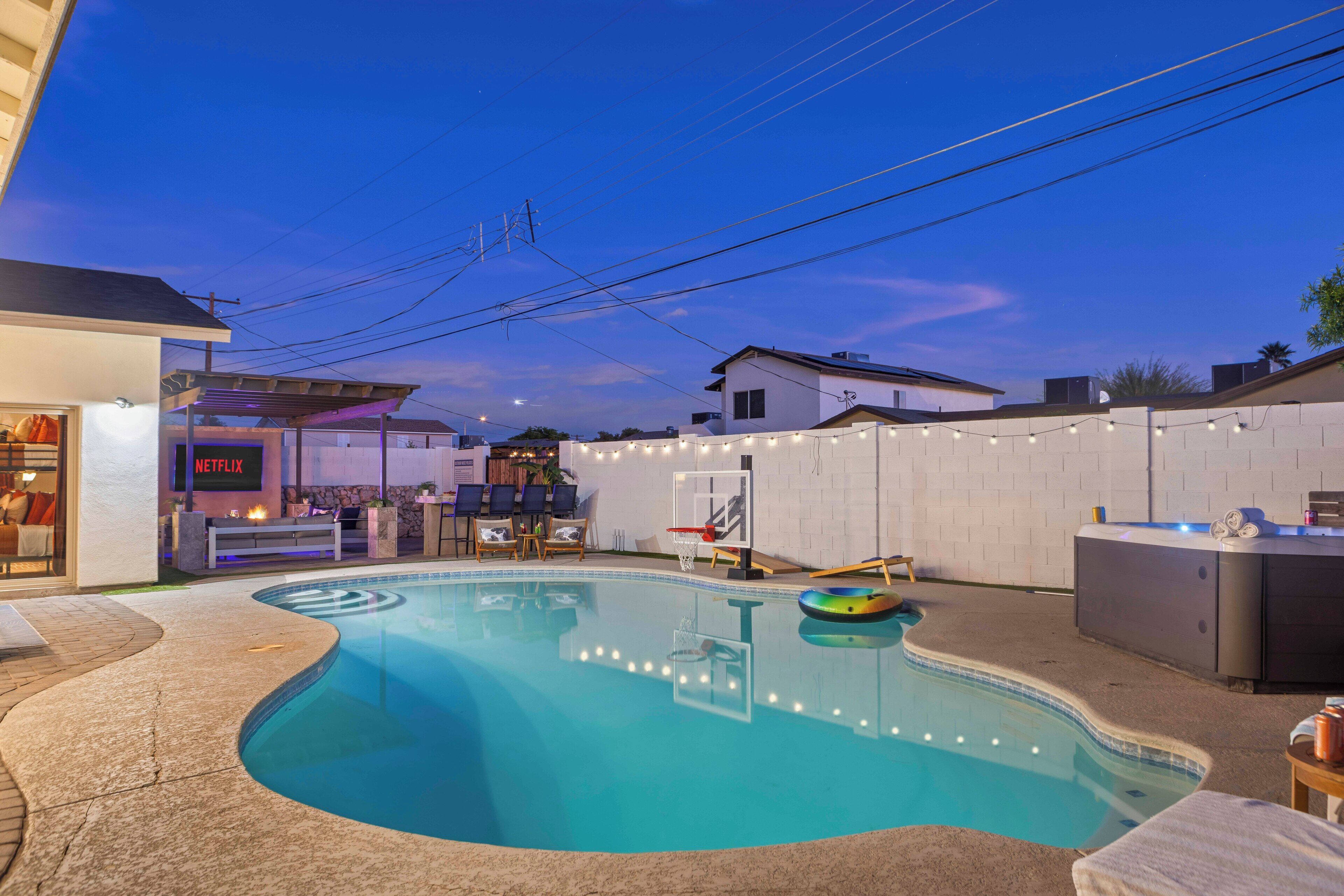 A swimming pool with a basketball hoop, lounge chairs, and a neon sign for a business.