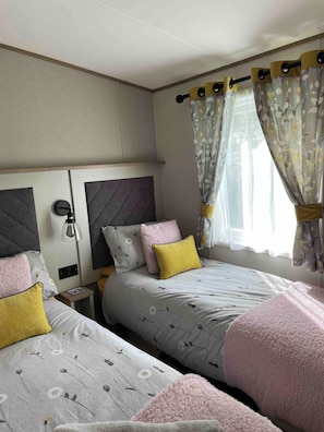 A bedroom with two beds, a nightstand, a lamp, and a window with curtains. - Charming Tattershall Mobile Home 3 Bedrooms, Private Entrance, Shared Pool (Lincoln)
