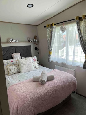 A neatly made bed with a pink fluffy blanket and rolled towels, a headboard with a 'Dream' sign, decorative pillows, and a window with curtains. - Charming Tattershall Mobile Home 3 Bedrooms, Private Entrance, Shared Pool (Lincoln)