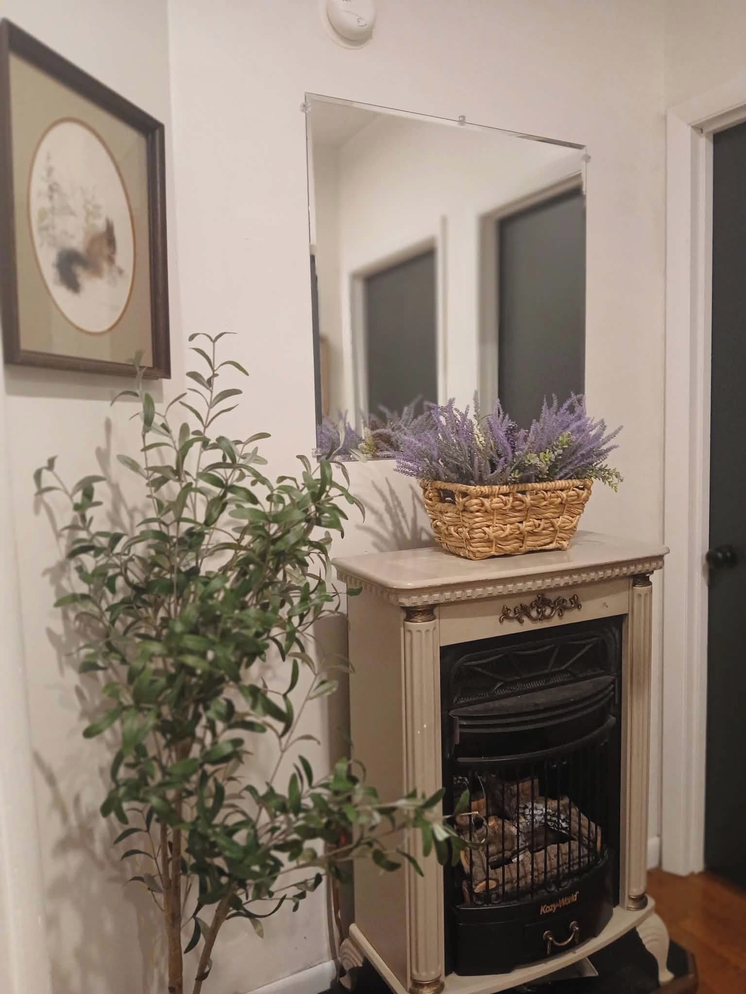 Une cheminée surmontée d’un panier tressé rempli de plants de lavande, un tableau encadré accroché au mur et une plante en pot placée à côté de la cheminée.