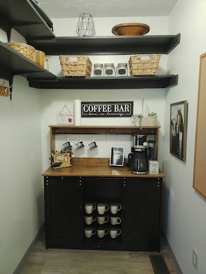 A coffee bar with a sign, a coffee maker, and mugs on shelves.