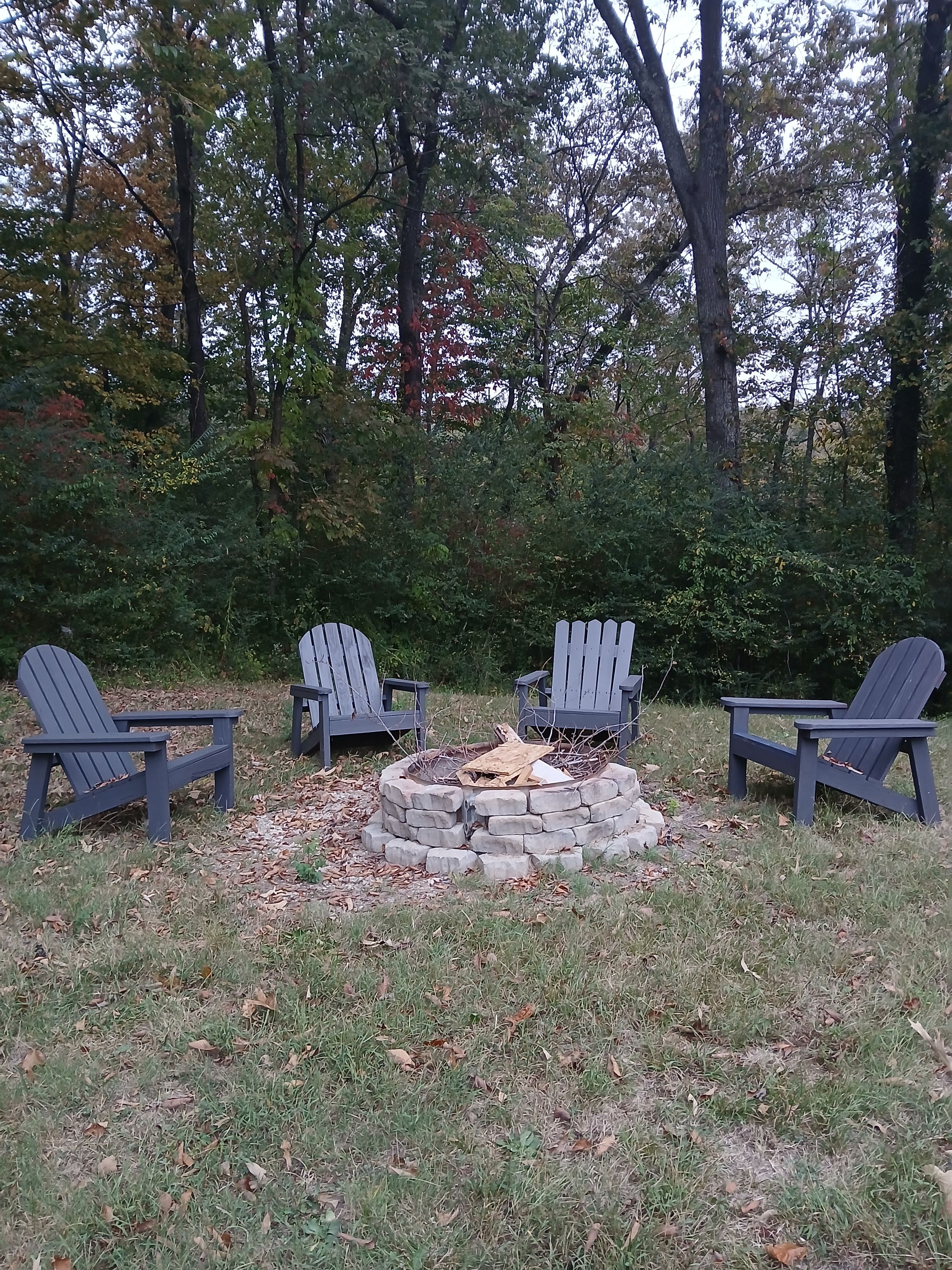 Un foyer extérieur entouré de quatre chaises Adirondack, le tout installé dans un espace verdoyant avec des arbres en arrière-plan.