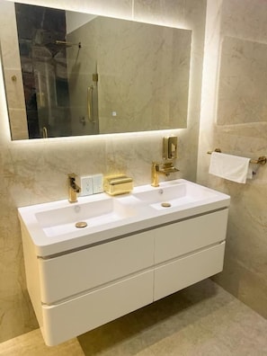 Casa de banho com toucador branco, duas pias, um espelho grande e um porta-toalhas.