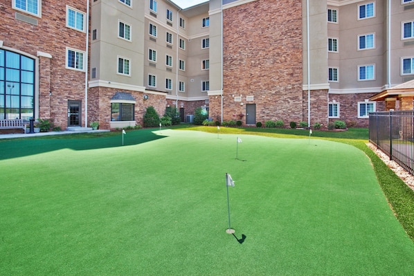 Un campo de golf con un putting green, rodeado por un edificio de varios pisos con paredes de ladrillo y color beige, ventanas grandes y una entrada con portón cerrado.