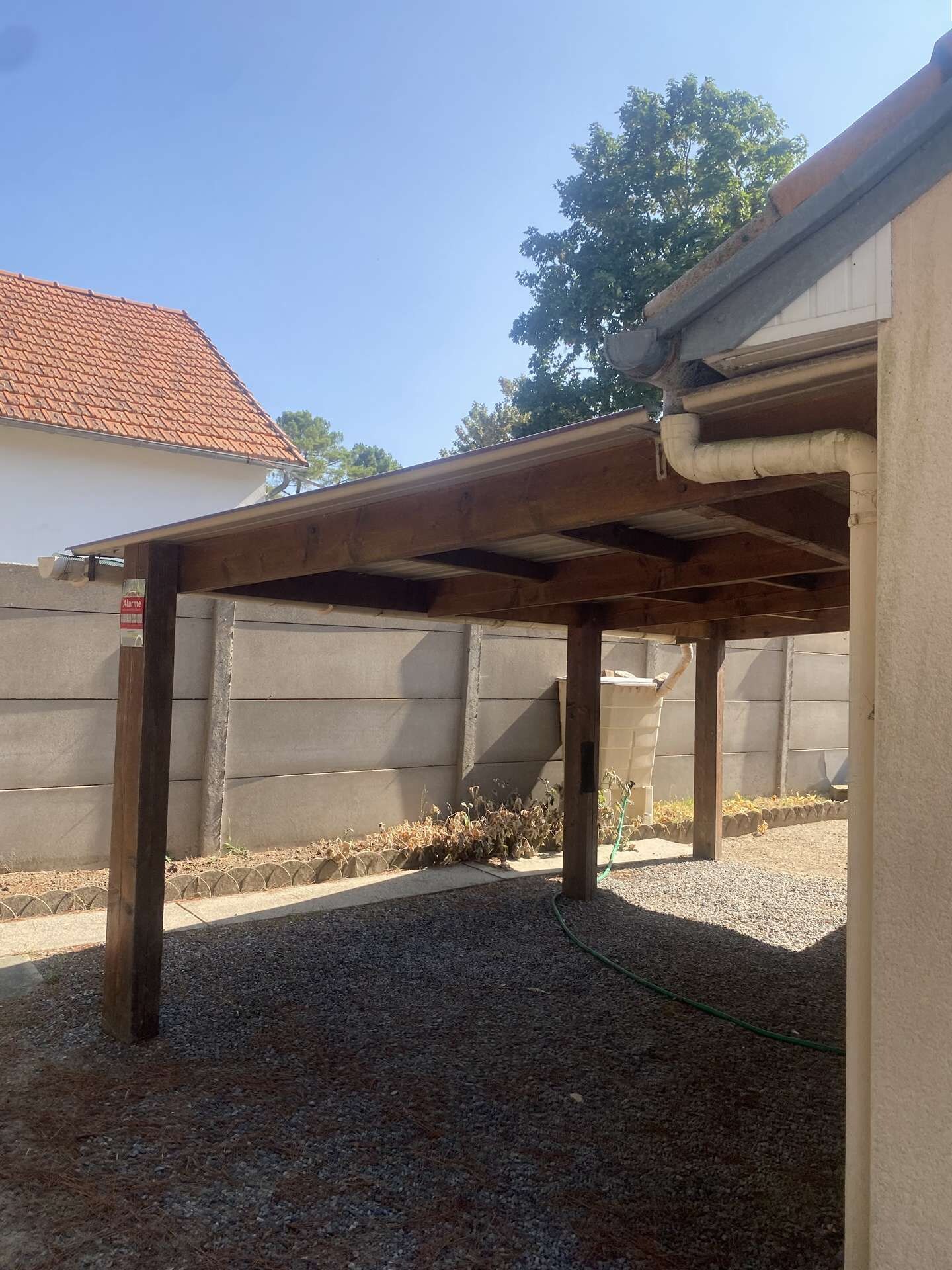 Un espace extérieur abrité, doté d’un sol en gravier, de poutres en bois et d’un tuyau d’arrosage.