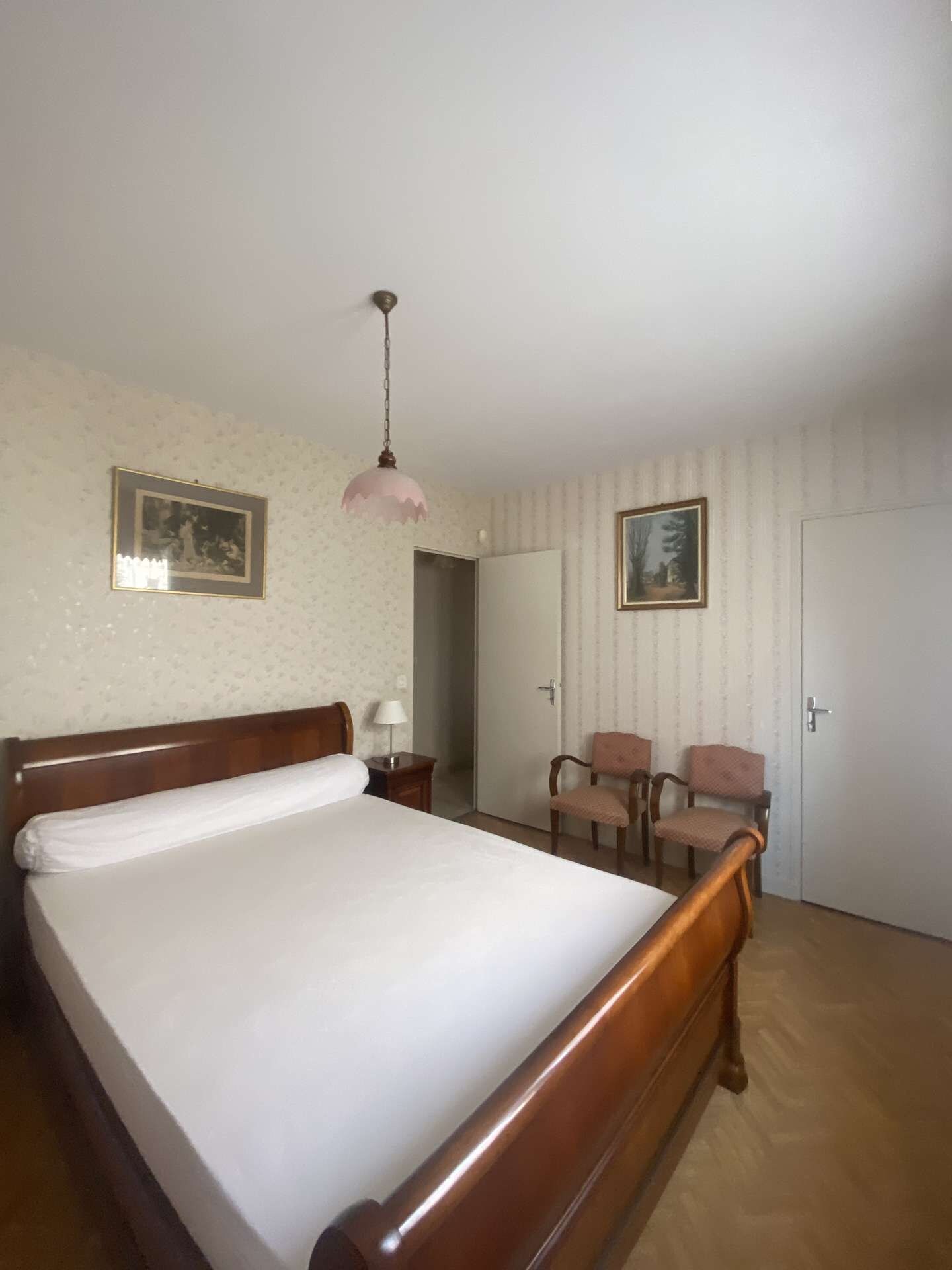 Une chambre avec un lit en bois, un luminaire suspendu, deux chaises et une porte.