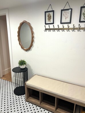 Ein Zimmer mit einem runden Spiegel, einem kleinen Tisch mit einer Pflanze, einer Bank und einem an der Wand befestigten Garderobenständer mit Haken.
