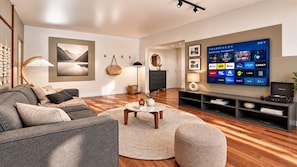 Ein modernes Wohnzimmer mit einem grauen Sofa, einem runden Couchtisch und einem Flachbildfernseher, der Streaming-Dienste anzeigt.