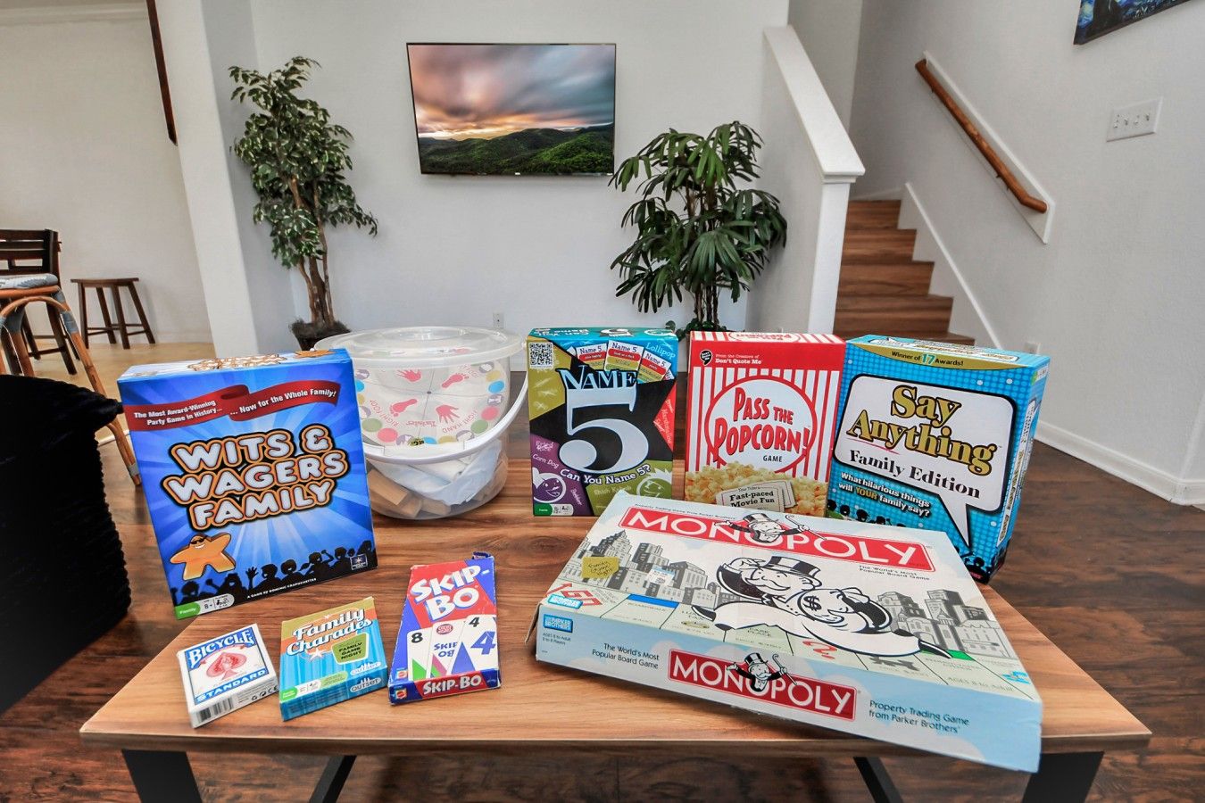A variety of board games on a wooden table in a room with a staircase and plants.