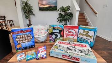 A variety of board games on a wooden table in a room with a staircase and plants.
