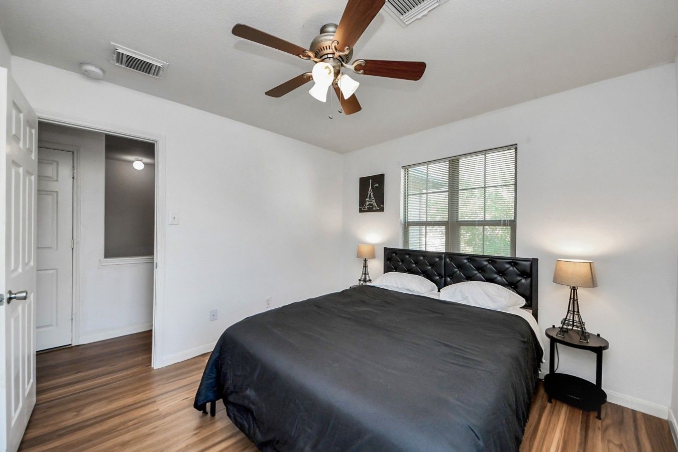 A bedroom with a bed, a ceiling fan, a window with blinds, and a door leading to another room.