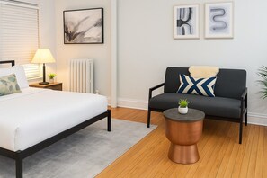 A modern bedroom with a bed, bedside table, lamp, and a grey sofa with a decorative pillow. - Stylish Studio near Boystown (Chicago)