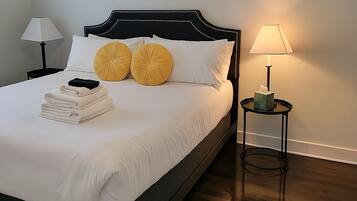 A neatly made bed with white linens, two decorative pillows, and a stack of folded towels. There are two bedside tables, one with a lamp and the other with a tissue box. The room has a dark wood floor and light-colored walls.