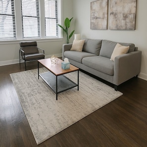 Ein Wohnzimmer mit grauem Sofa, einem Holztisch, einem schwarzen Sessel und einem großen Fenster mit Jalousien.