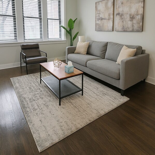 Una sala de estar con un sofá gris, una mesa de centro de madera, una silla negra y un ventanal con persianas.